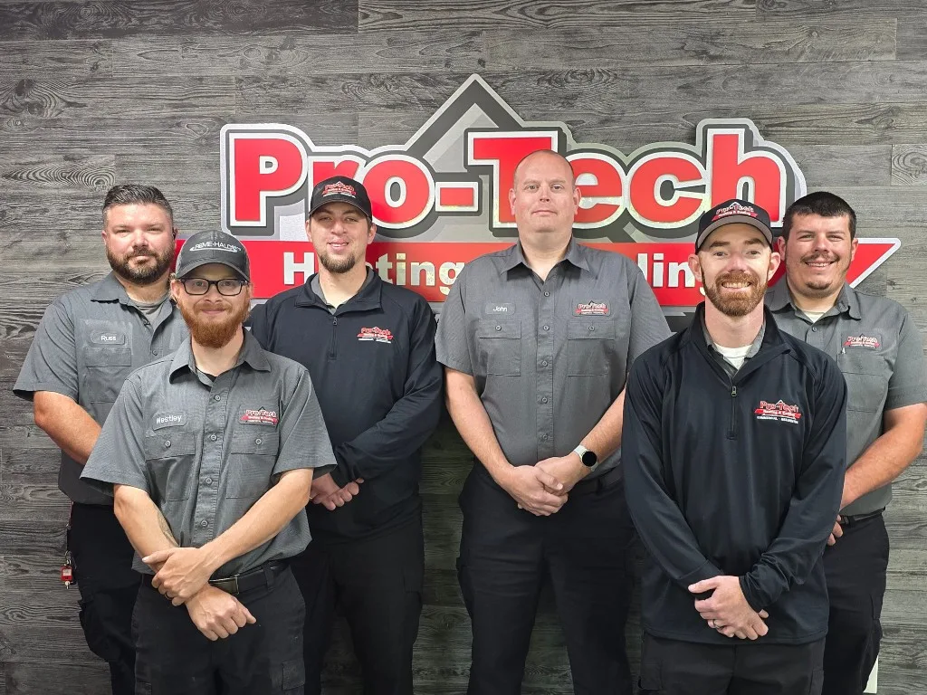 Pro-Tech Heating & Cooling certified HVAC service technicians in front of the Pro-Tech logo wall at our Grand Rapids, MI shop